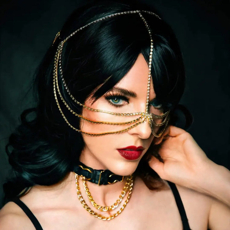 Glamorous portrait featuring dark hair, red lips, gold chain accessories, and a delicate face chain draping across one eye.