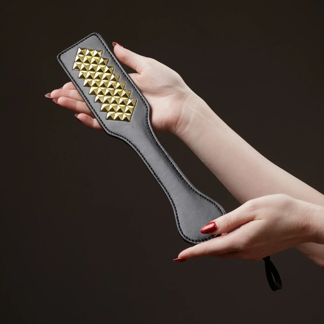 Hand holding a black spanking paddle with gold pyramid studs on a dark background