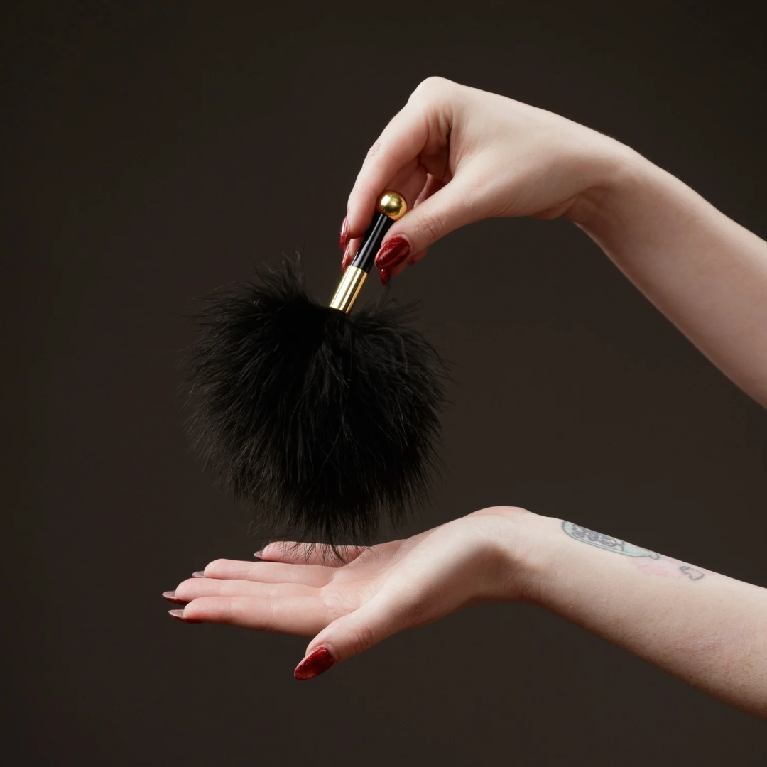 Black fluffy small tickler with gold handle held by a hand against a dark background