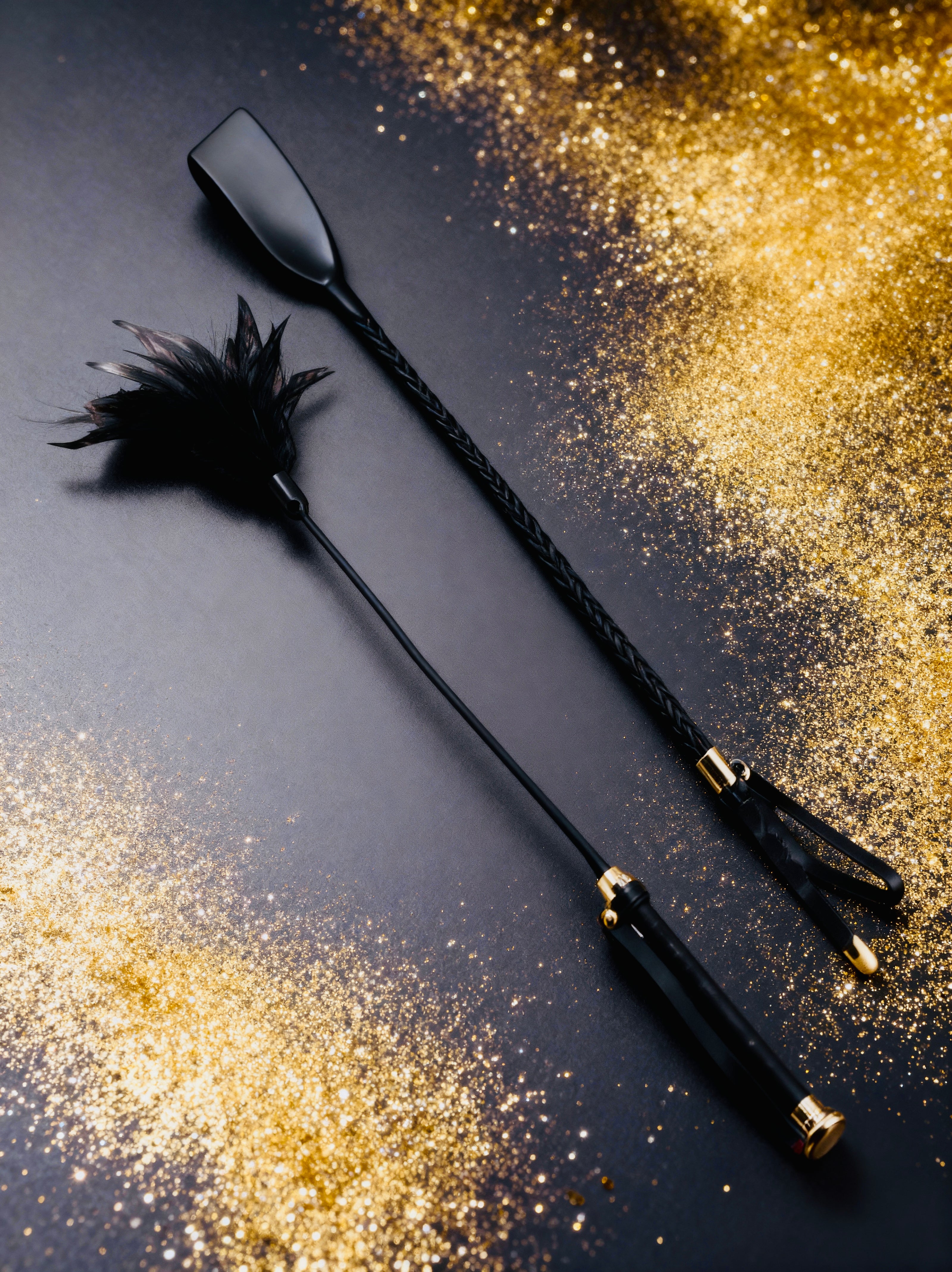 Black bondage feather tickler and playful spanking whip set on a dark surface with gold glitter
