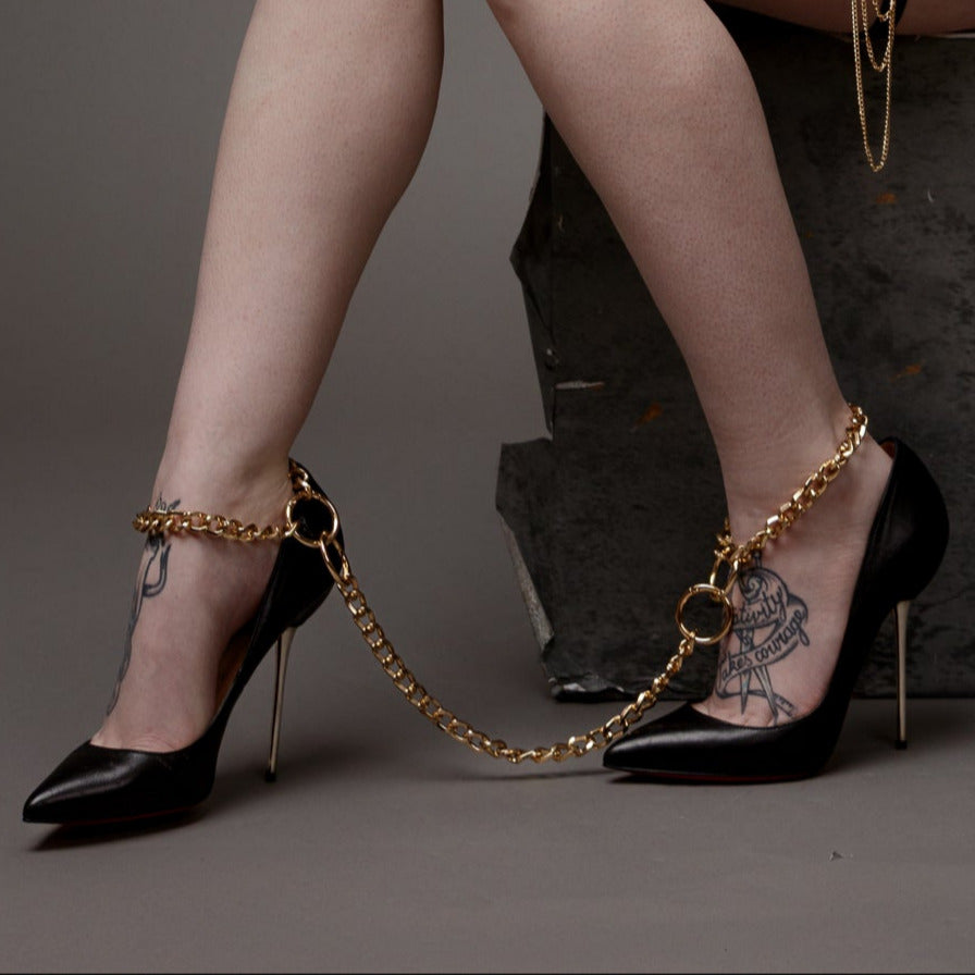 Black high-heeled shoes with gold kink chain ankle straps on a neutral background