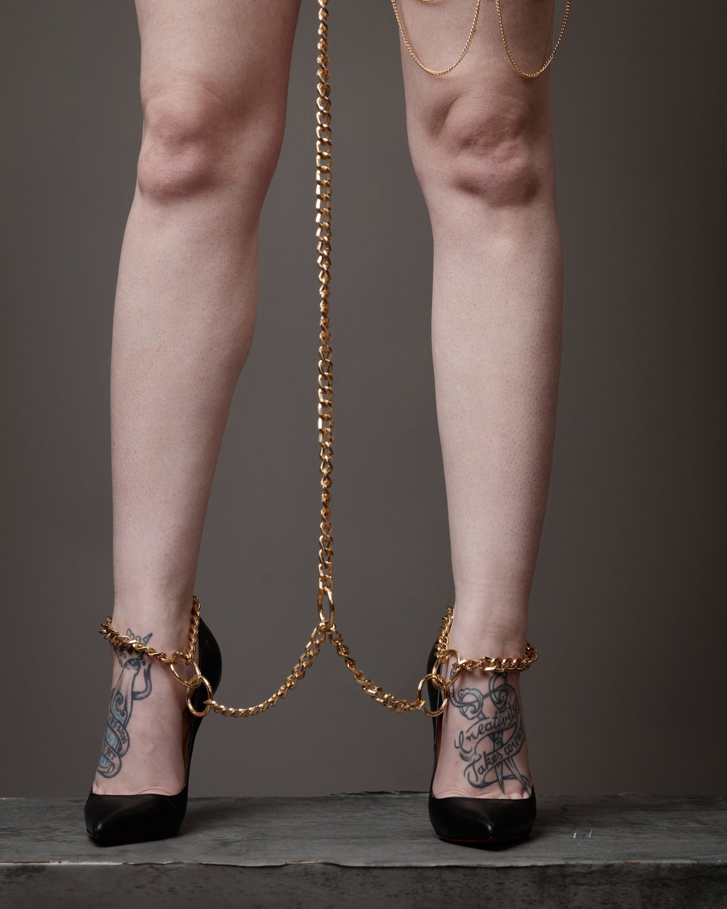 Legs wearing high heels with gold chains and tattoos on a neutral background