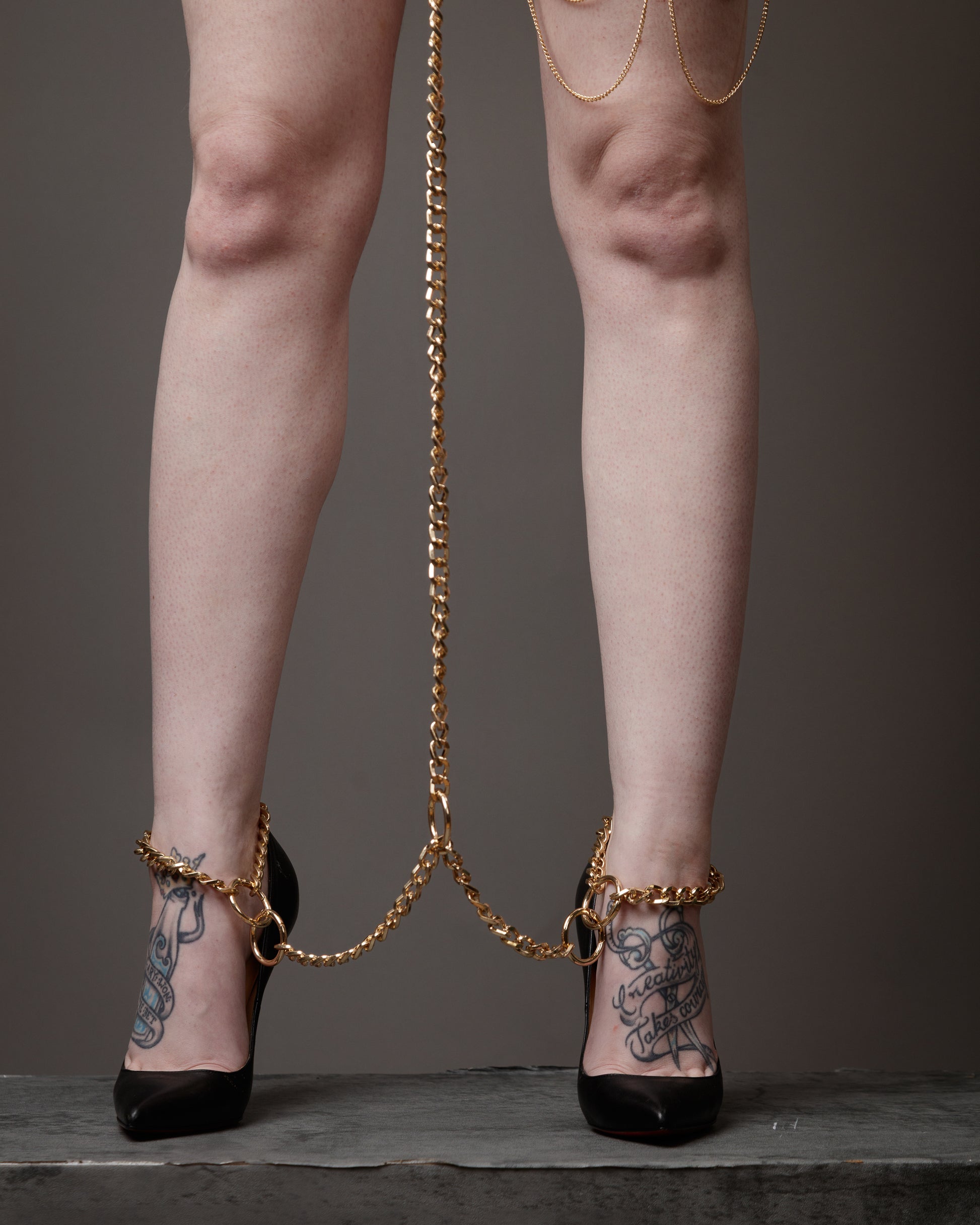 Legs wearing high heels with gold chains and tattoos on a neutral background