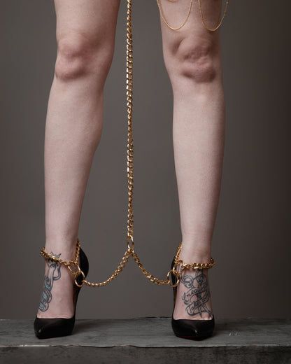 Legs wearing high heels with gold chains and tattoos on a neutral background